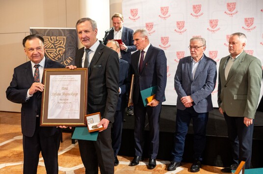 Mężczyzna trzymający dyplom i medal, otoczony przez grupę elegancko ubranych mężczyzn, stoi na tle ścianki z logo.