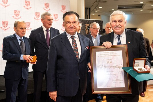 Kilku mężczyzn na oficjalnej ceremonii. Jeden trzyma oprawiony dyplom, drugi otwarte pudełko z medalem. W tle widoczne logo Mazowsza.