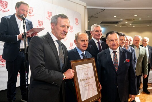 Grupa elegancko ubranych mężczyzn pozuje do zdjęcia na uroczystości. Jeden z nich trzyma dużą ramę z nagrodą dla Henryka Apostela. Stoją na tle ścianki z logo Mazowieckiego ZPN.