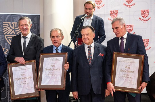 Czterech mężczyzn w garniturach stoi obok siebie, trzymając duże, oprawione dyplomy. Za nimi mężczyzna z mikrofonem. W tle widoczne jest logo 