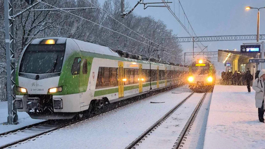 Zasypana śniegiem stacja kolejowa; widoczne dwa pociągi na torach i pasażerowie w płaszczach stojący na peronie.