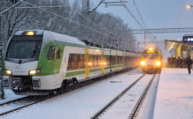 Zasypana śniegiem stacja kolejowa; widoczne dwa pociągi na torach i pasażerowie w płaszczach stojący na peronie.