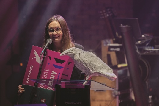 Uśmiechnięta osoba z nagrodą przy mikrofonie na scenie.