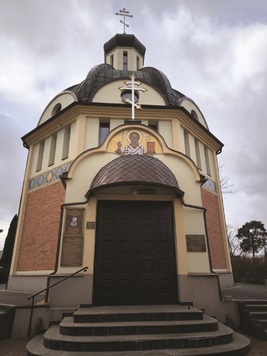 Cerkiew z kopułą i krzyżem, zdobiony ikoną nad drzwiami. Schody prowadzą do drewnianych drzwi. Ściany z jasnego tynku i cegły.