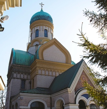 Cerkiew z zielonymi kopułami, zdobiona detalami architektonicznymi, otoczona zielenią na tle jasnego nieba.