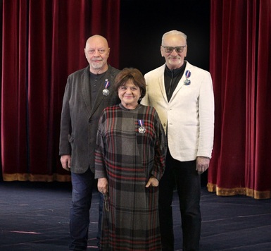 Trzy osoby stoją na tle czerwonej kurtyny teatralnej. Każda z nich ma przypięty medal na piersi.