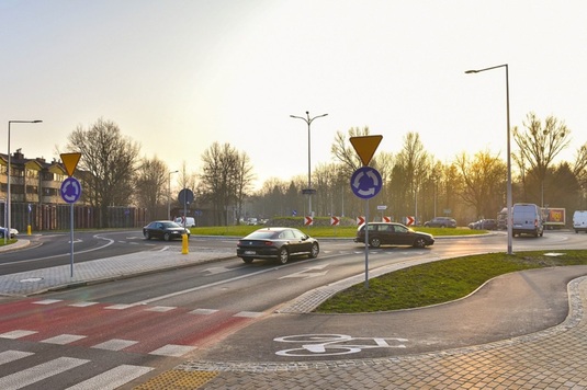 Rondo drogowe o poranku z samochodami poruszającymi się po jezdni. Znaki drogowe i ścieżka rowerowa na pierwszym planie, drzewa wokół drogi.