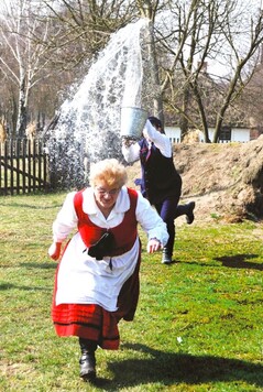 Osoba w tradycyjnym stroju wiejskim ucieka przed wodą wylewaną z wiadra przez inną osobę. W tle widać drzewa i płot.
