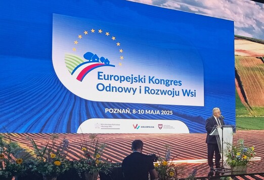 Mężczyzna przemawia na tle dużego ekranu z logo Europejskiego Kongresu Odnowy i Rozwoju Wsi. Obok bukiety kwiatów, a za nim malowniczy widok pól. Wydarzenie ma miejsce w Poznaniu w maju 2025 roku.
