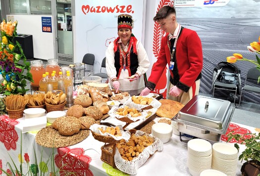 Stół z regionalnymi potrawami, napojami i pieczywem, przy którym stoją dwie osoby w tradycyjnych strojach. W tle baner z napisem 