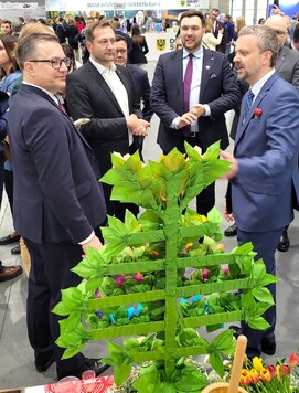 Grupa mężczyzn w garniturach rozmawia przy stoisku z kolorowymi dekoracjami z papierowych kwiatów i zielonych liści. W tle widać innych uczestników wydarzenia.