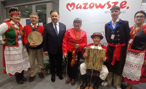Grupa ludzi ubrana w tradycyjne stroje ludowe, stojąca przed białą ścianą z napisem 
