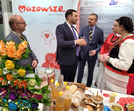 Grupa osób rozmawia przy stole z regionalnymi produktami spożywczymi, na tle stoiska promującego Mazowsze. Dwie osoby są w tradycyjnych strojach ludowych, a pozostałe w garniturach. Obok stoi wazon z kolorowymi kwiatami.