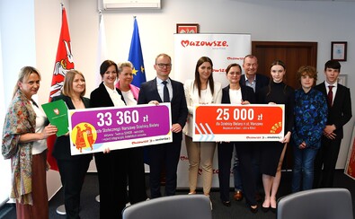 Grupa osób stoi razem, trzymając duże tablice z kwotami 33 360 zł i 25 000 zł. W tle widoczne są flagi i baner Mazowsze.