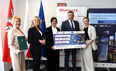 Grupa osób stoi z dużym czekiem na kwotę 451 360 zł na tle flag i banerów Mazowsza, związanych z funduszami europejskimi.