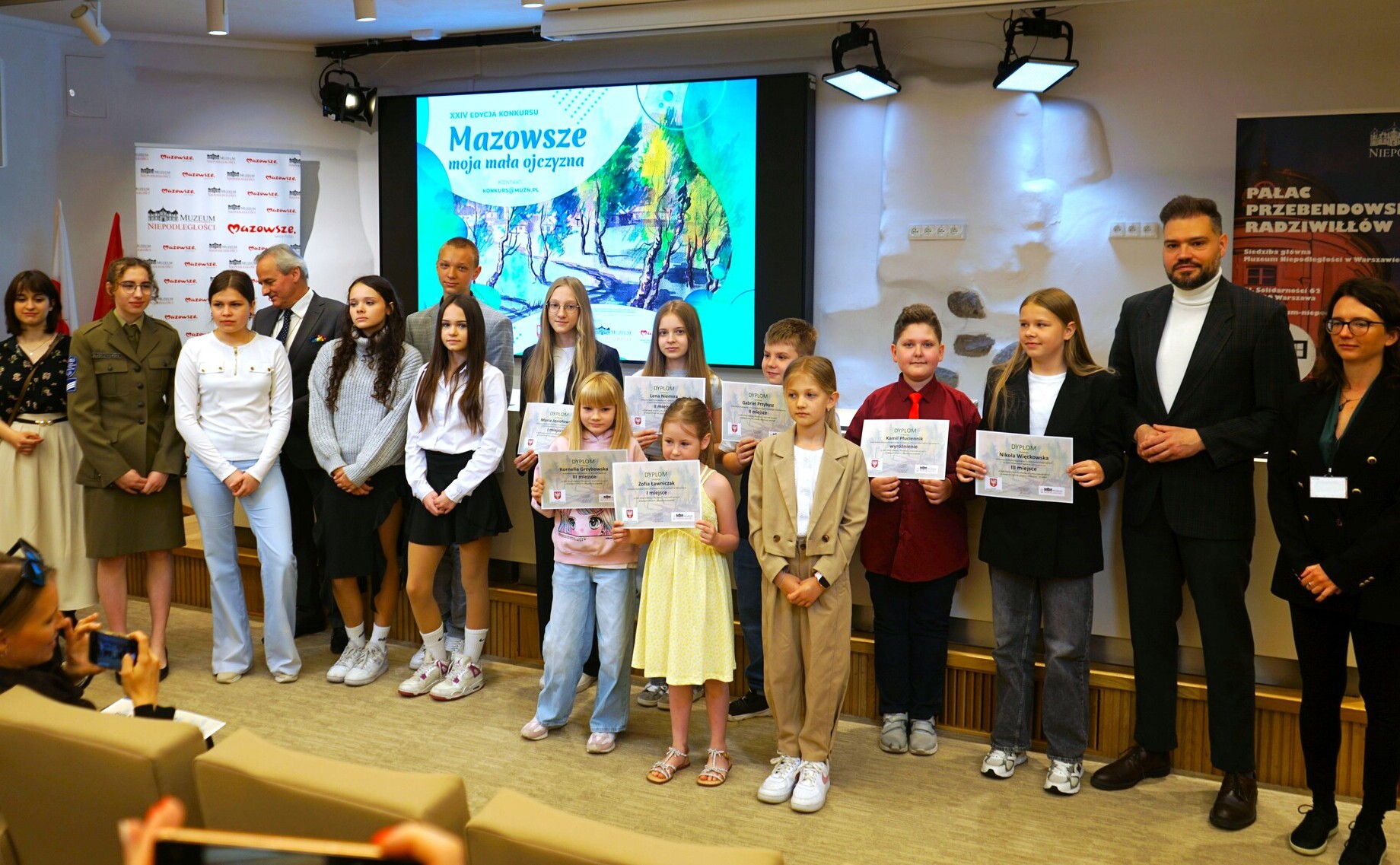 Grupa ludzi pozuje do zdjęcia podczas uroczystości rozdania dyplomów w sali konferencyjnej. W tle widać duży ekran z napisem „Mazowsze moja mała ojczyzna”.