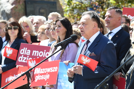 Grupa osób stoi na dworze, jedna z nich przemawia do mikrofonu. Wiele osób trzyma czerwone tablice z białymi napisami i symbolami.