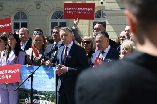 Grupa osób w garniturach stoi przed budynkiem z oknami łukowymi. Jeden mężczyzna przemawia do mikrofonu. Inni trzymają czerwone tabliczki z napisami, a w tle widać transparent promujący głosowanie.