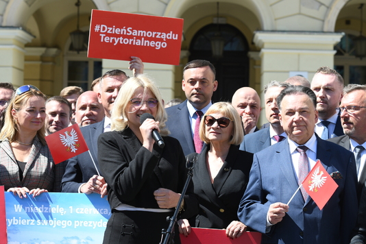 Grupa osób w eleganckich ubraniach stoi na zewnątrz przed budynkiem. Kobieta z blond włosami mówi do mikrofonu. Widać flagi z orłem i transparent z napisem 