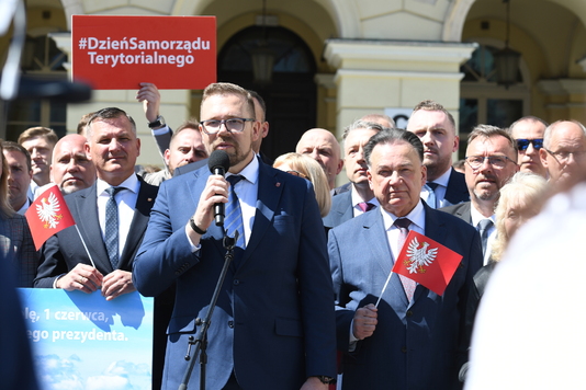 Grupa ludzi w garniturach podczas wydarzenia na świeżym powietrzu, jedna osoba przemawia do mikrofonu. W tle widoczny transparent z napisem 