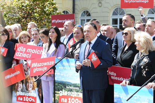Grupa ludzi w eleganckich strojach stoi na zewnątrz z różnymi transparentami, m.in. 