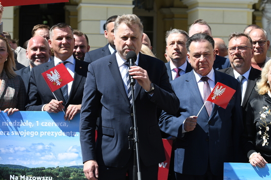 Grupa elegancko ubranych osób stoi na tle budynku. Mężczyzna na pierwszym planie mówi do mikrofonu, inni trzymają czerwone flagi z białym orłem.